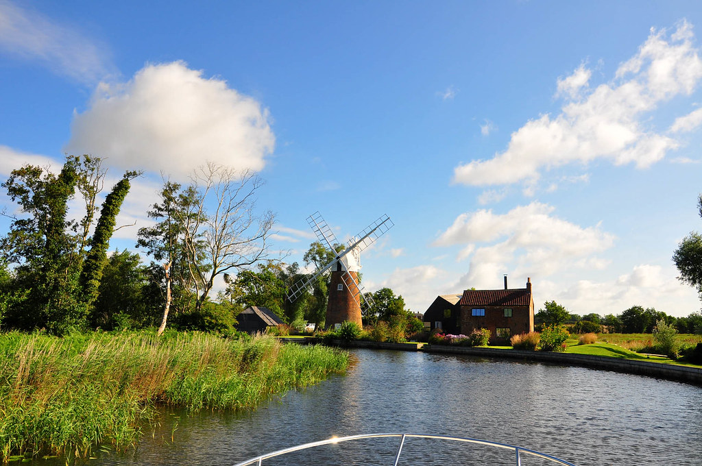 Photo of part of the Broads taken from the front of  a boat showing a windmills and a house. 