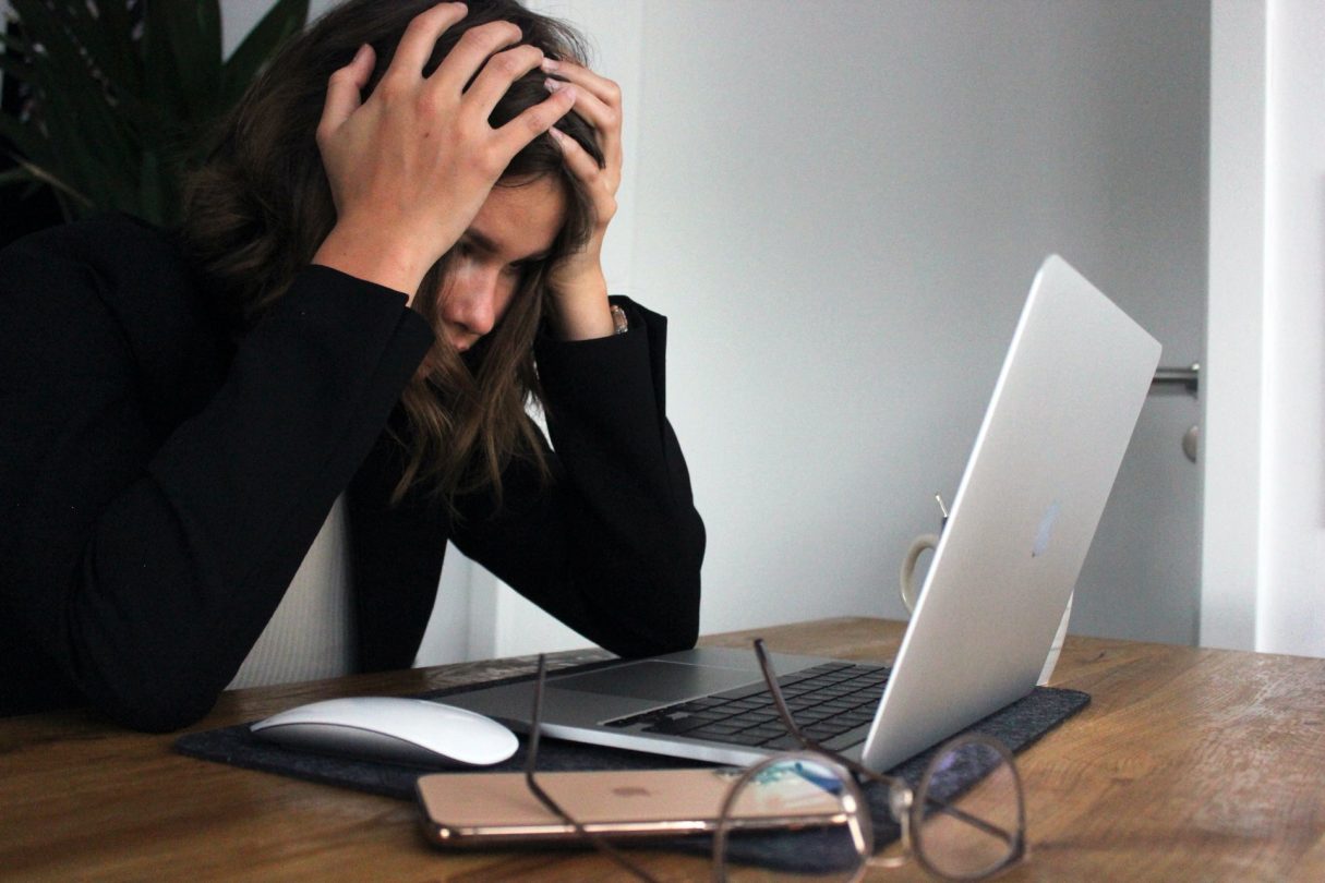 A lady sitting at a laptop appearing stressed with her hands on her head. 